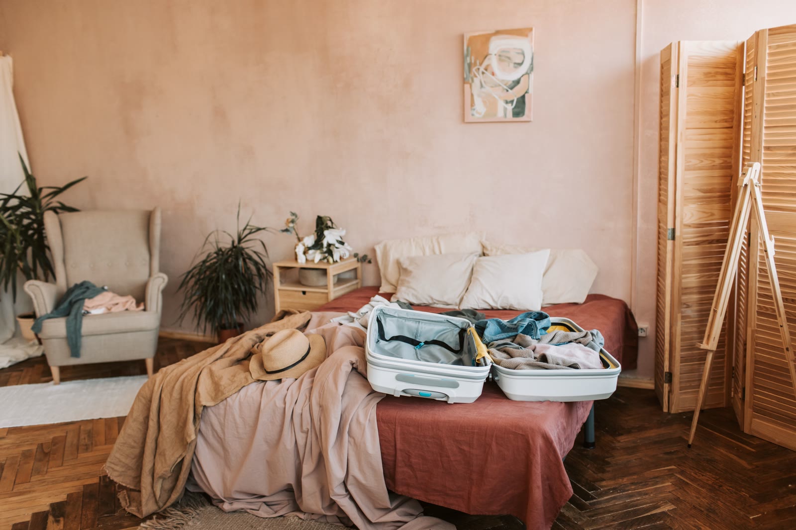 Maleta abierta a medio hacer sobre una cama deshecha, con un sombrero de paja al lado, en un dormitorio de paredes rosas