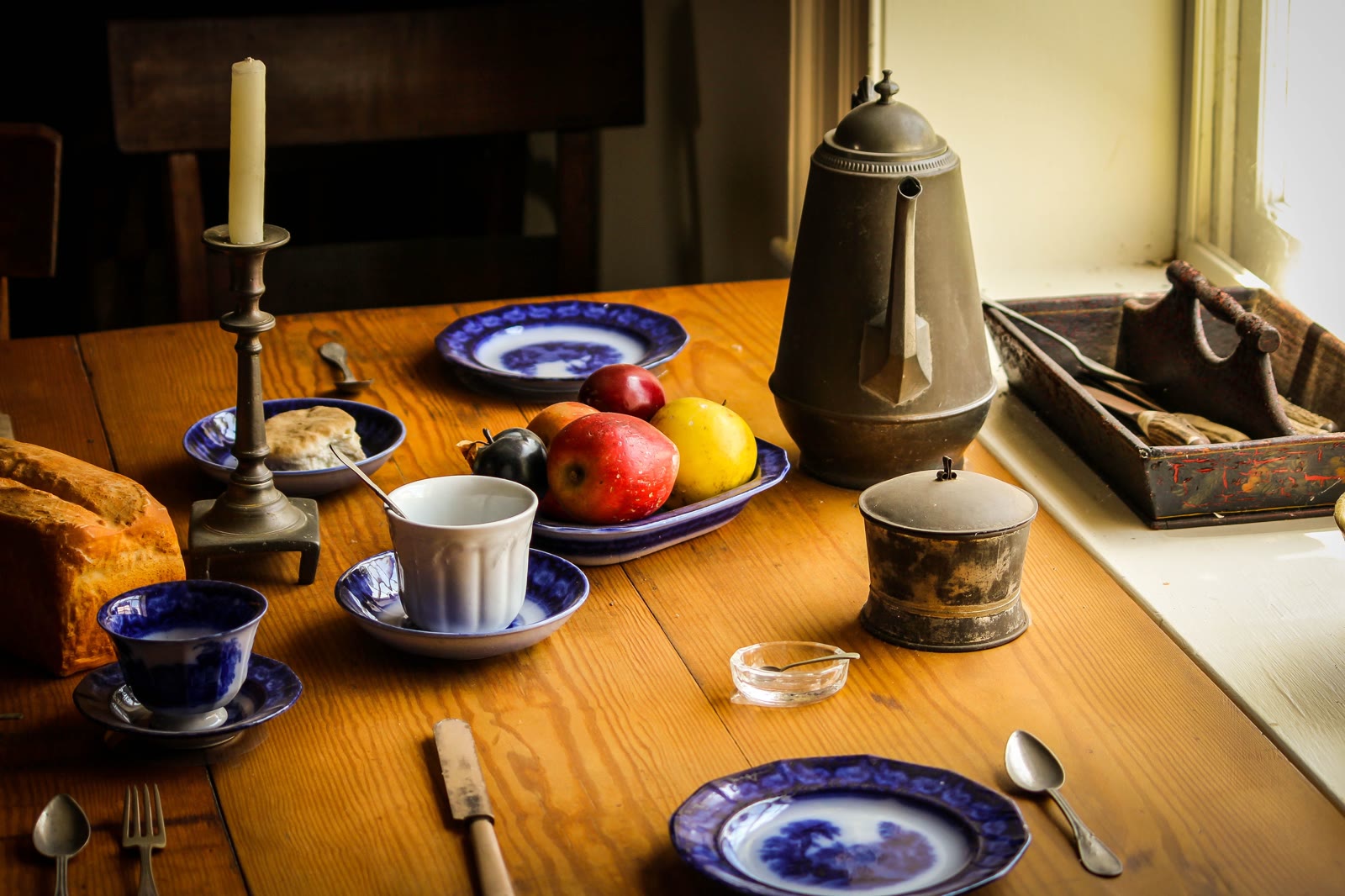 Mesa de madera antigua puesta junto a una ventana, con vajilla azul, pan, fruta y una cafetera de metal