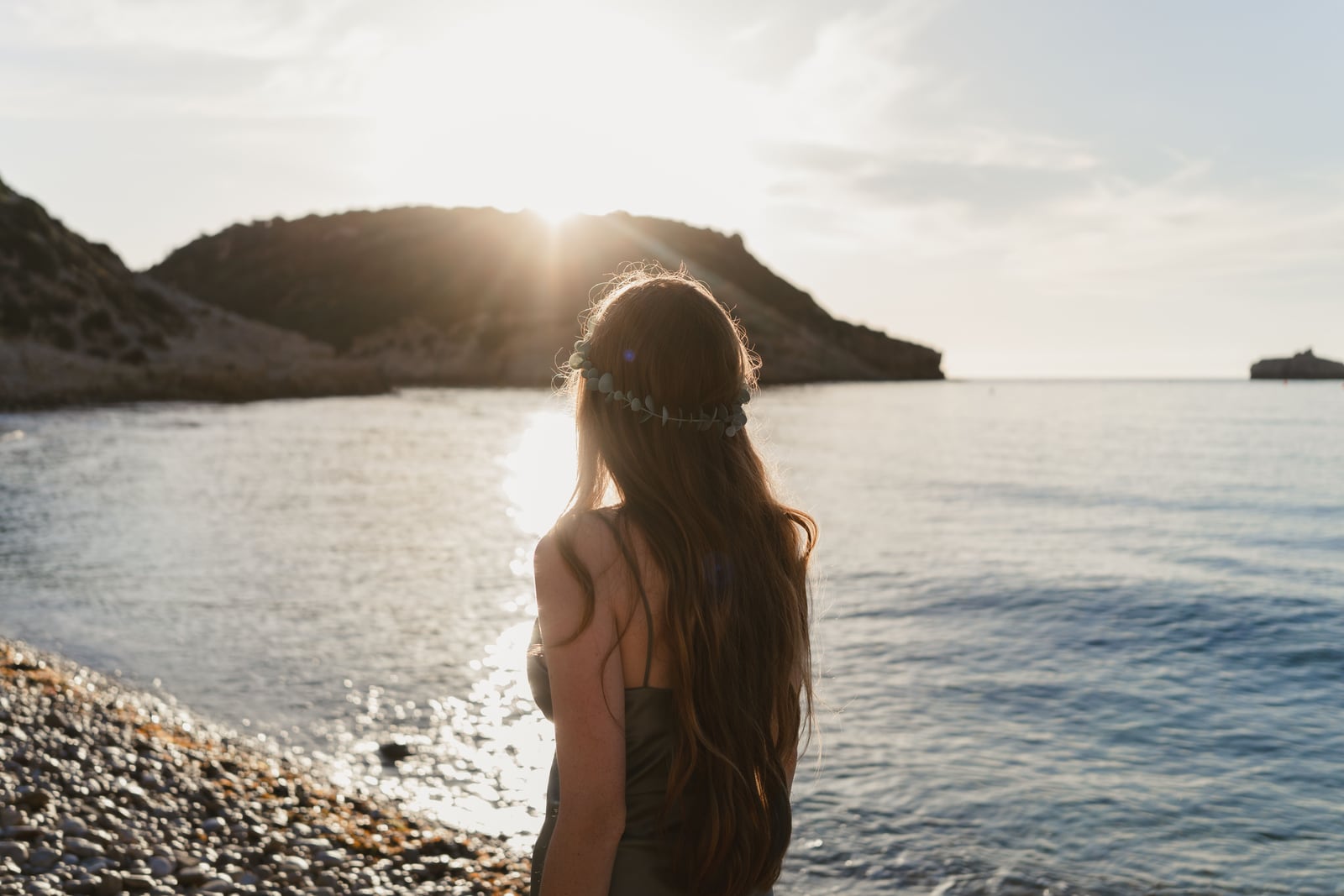 Mujer de espaldas, de pie en la orilla, mirando el mar a contraluz al atardecer